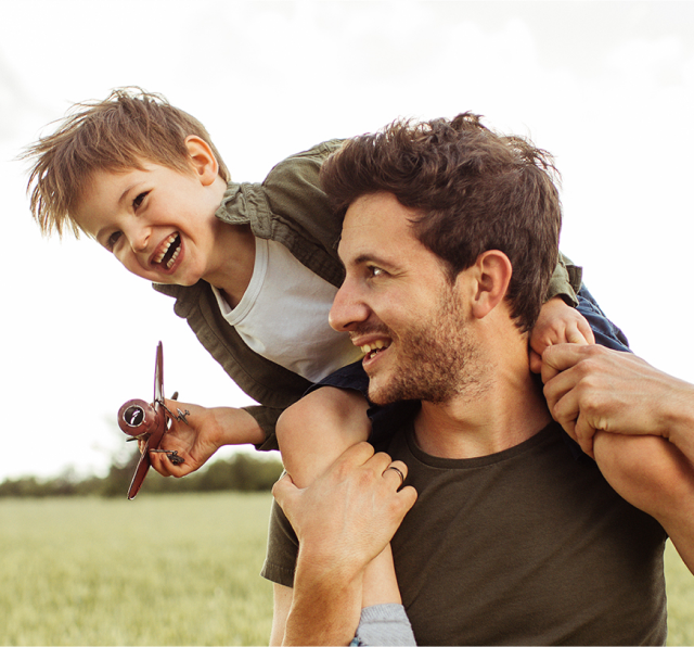 Happy son rides on dad's shoulders outside in green field
